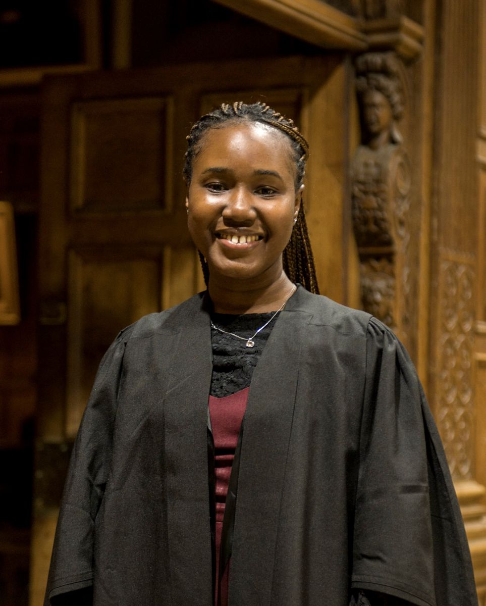 Portrait of Paula-Peace James-Okoro wearing an academic gown. She has brown skin and black hair and is smiling.